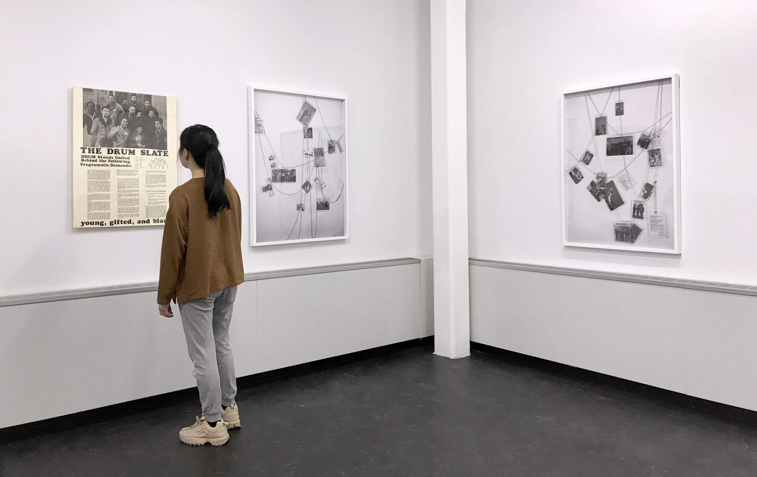 Photo of an exhibition showing a white table with archive items in the foreground. Hanging on the two rear walls are art objects: on the left wall there is a poster of Detroit’s Revolutionary Union Movement’s Slate, and hanging on the right wall is a sculpture made of many laminated images related to Detroit’s Revolutionary Black Workers Movement (1960s-1970s) all linked together by a beaded chain and arranged rather randomly