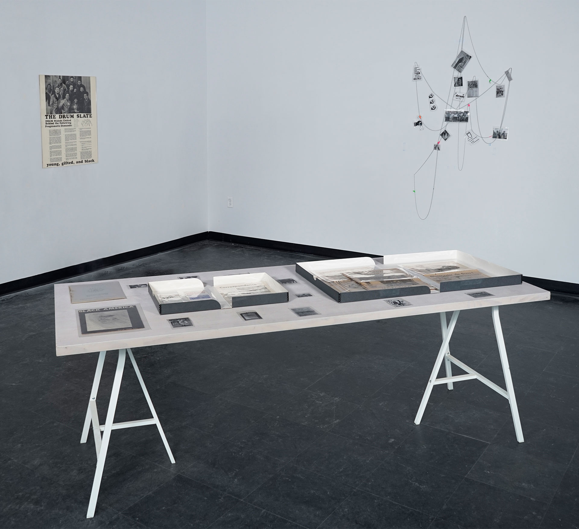 Photo of an exhibition showing a white table with archive items in the foreground. Hanging on the two rear walls are art objects: on the left wall there is a poster of Detroit’s Revolutionary Union Movement’s Slate, and hanging on the right wall is a sculpture made of many laminated images related to Detroit’s Revolutionary Black Workers Movement (1960s-1970s) all linked together by a beaded chain and arranged rather randomly